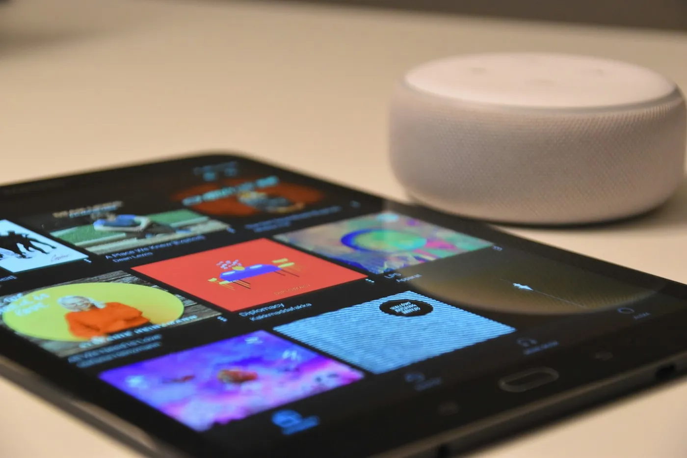 A tablet showing a media app grid next to a fabric-top smart speaker on a table