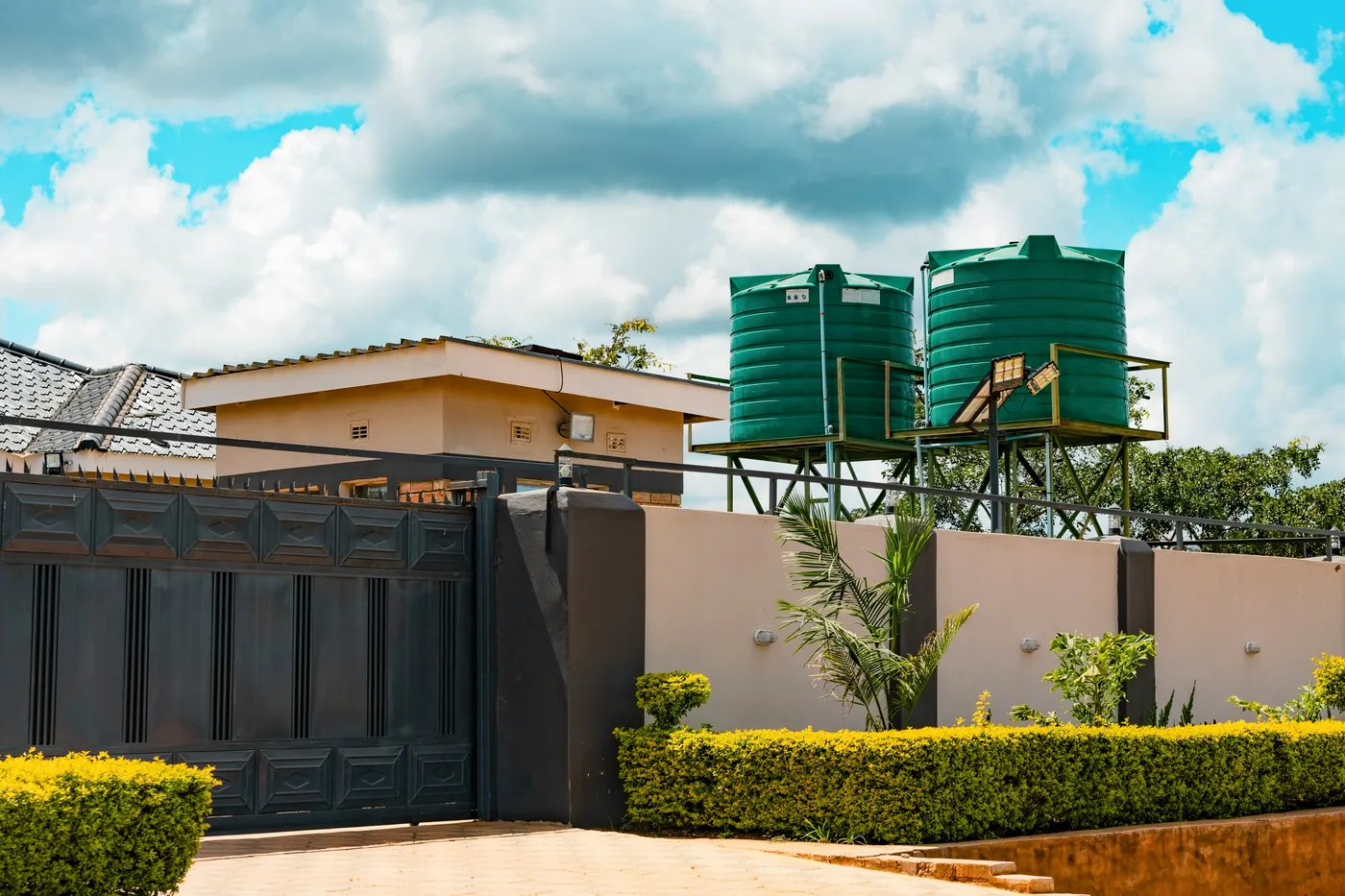 Dark metal gate set in a beige rendered boundary wall, with a trimmed hedge in front and water tanks on stands behind