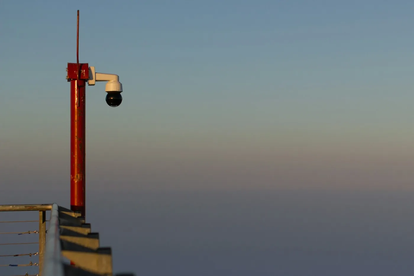 A PTZ dome camera on a weathered reddish pole overlooking the sea at dusk