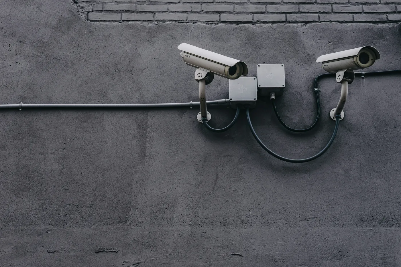 Two wired bullet CCTV cameras on a grey rendered wall, joined by conduit to two junction boxes