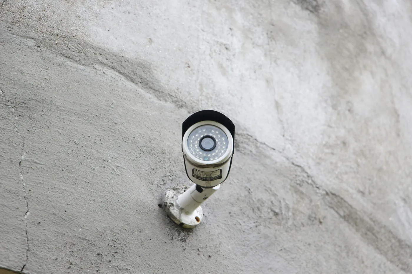 White bullet CCTV camera with a short hood, on a bracket on a pale rendered wall in daylight