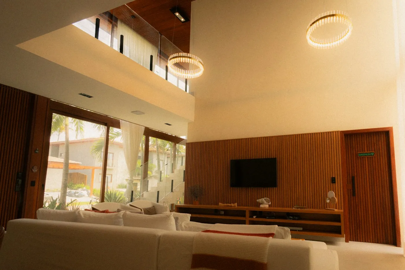 Double height villa living room with warm glowing ring pendant lights and palm trees visible outside