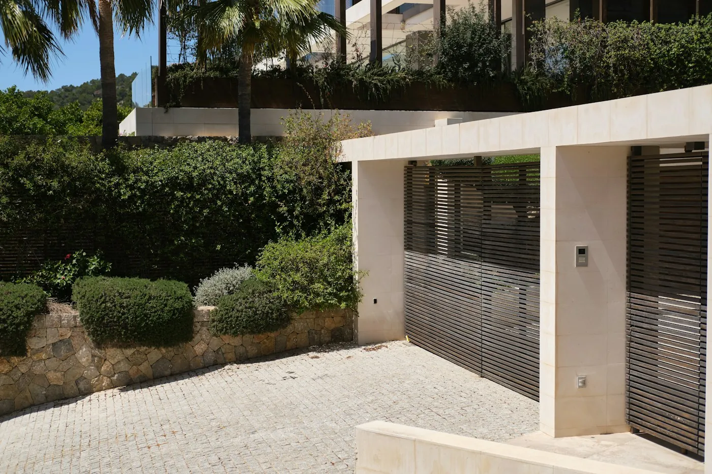 Modern slatted villa gate with stone pillars and a wall mounted intercom panel beside palm trees