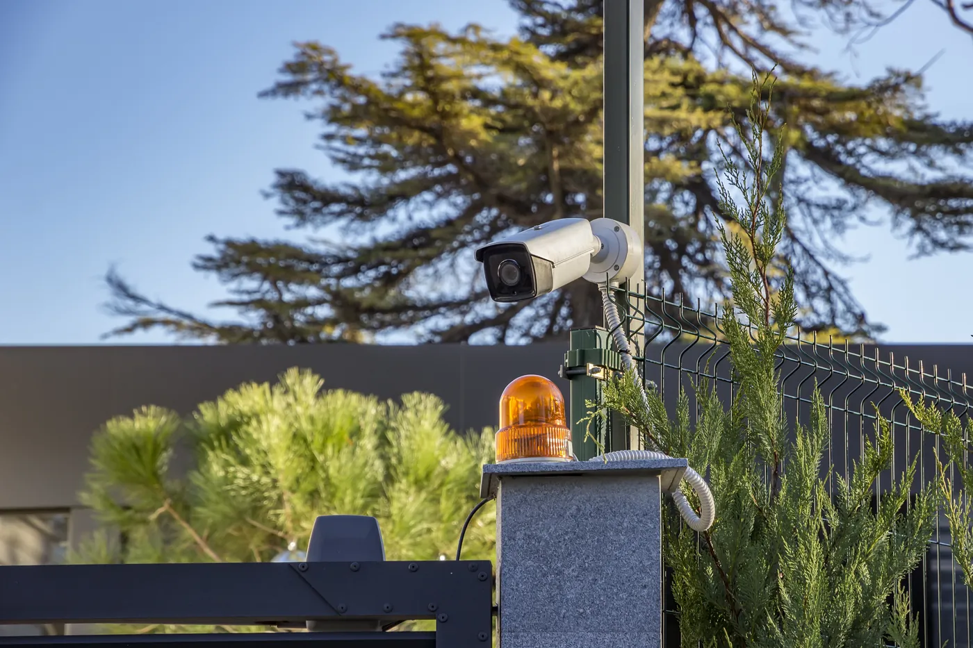 Outdoor security camera on a pole beside a villa garden wall in bright sun