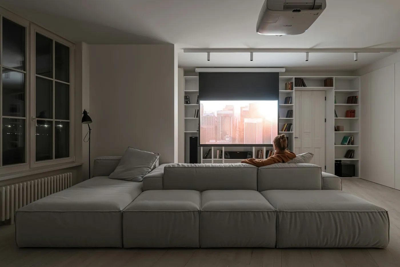 A person on a large grey sectional sofa watching a projector screen in an upscale media room at night