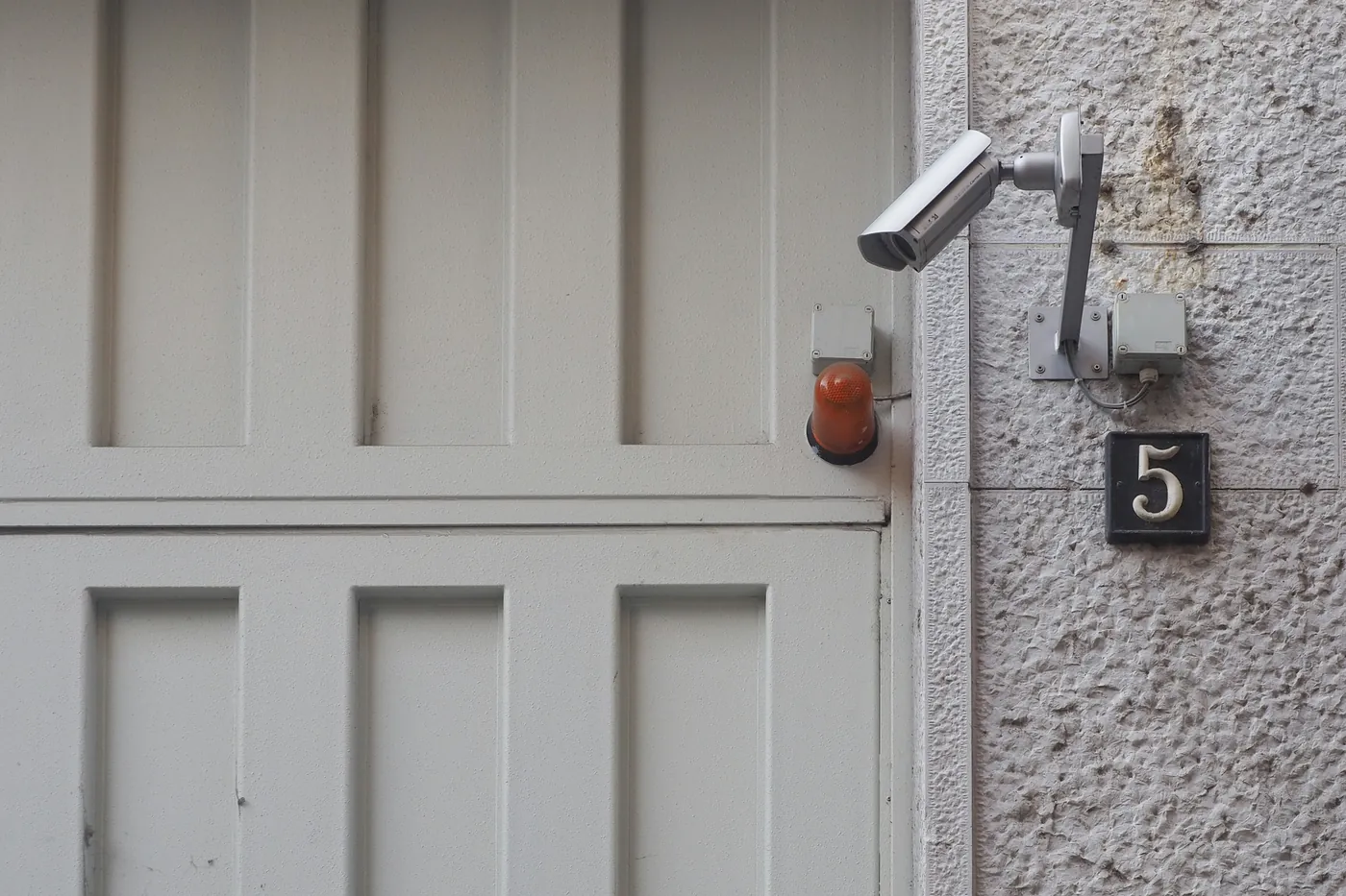 Pale panelled door with an orange alarm beacon beside it and a grey wired security camera on a bracket above a house number on a textured wall