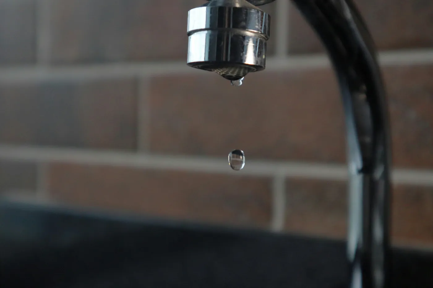 A single water drop falling from a chrome tap against dark brick, the kind of slow water leak that is easy to miss
